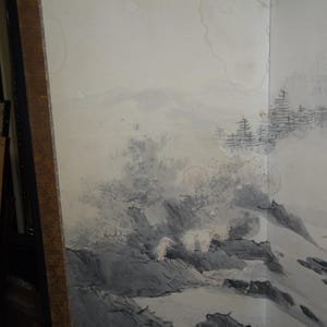 May include: A framed, multi-panel artwork featuring a landscape scene. The artwork depicts a mountainous terrain with trees and water, rendered in shades of gray and white. The frame is a dark brown color.