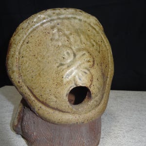 May include: A ceramic sculpture of a tanuki, a Japanese raccoon dog. The sculpture has a round head with a large opening and a textured body. The tanuki is a traditional figure in Japanese folklore, often associated with good fortune and prosperity.