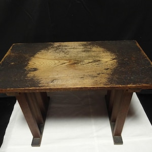Puede incluir: Una pequeña mesa rectangular de madera con un acabado envejecido. La superficie es principalmente marrón claro con bordes marrón oscuro. Las patas son de un marrón más oscuro y están anguladas para dar soporte. La mesa mide unos 61 cm de largo, 41 cm de ancho y 30 cm de alto.