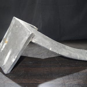 Puede incluir: Una cabeza de hacha de metal gris desgastado con un mango curvo. La cabeza del hacha es rectangular con un borde afilado y muestra signos de desgaste. El mango se extiende desde la cabeza, curvándose suavemente hacia un extremo redondeado. El fondo es oscuro.