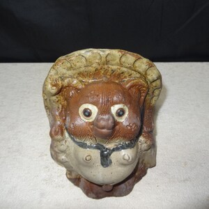 May include: A ceramic Tanuki statue, a traditional Japanese raccoon dog figure. The statue features a brown body, a beige hat, and a white belly. It has large, round eyes and a smiling expression. The figure is a symbol of good fortune.