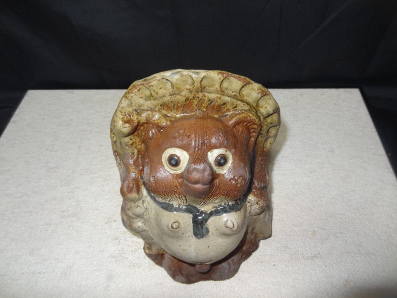 May include: A ceramic Tanuki statue, a traditional Japanese raccoon dog figure. The statue features a brown body, a beige hat, and a white belly. It has large, round eyes and a smiling expression. The figure is a symbol of good fortune.
