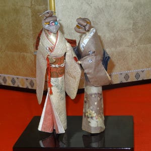 以下が含まれることがあります： 伝統的な着物を着た2体の日本の紙人形。左の人形は、花柄と赤いアクセントが付いたクリーム色の着物を着ています。右の人形は茶色の着物を着ています。どちらの人形も黒い台座の上に立っています。