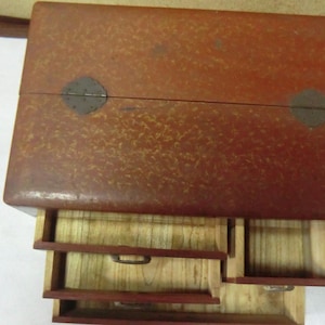 May include: A wooden box with a red finish and a speckled pattern. The box has three drawers, each with a different size. The top of the box has two metal hinges.