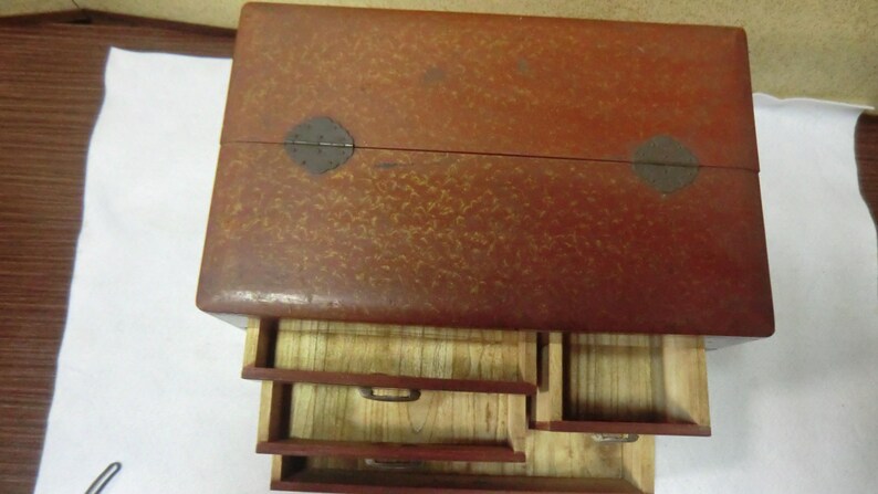 May include: A wooden box with a red finish and a speckled pattern. The box has three drawers, each with a different size. The top of the box has two metal hinges.