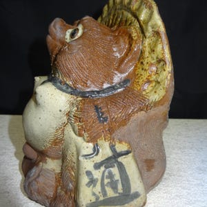 May include: A ceramic sculpture of a tanuki, a Japanese raccoon dog. The sculpture is primarily brown and tan with a black collar and a sack with Japanese characters. The tanuki has a whimsical expression and a textured surface.
