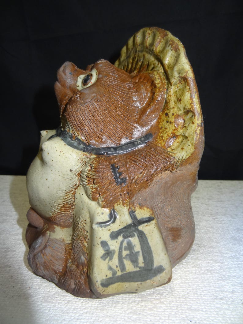 May include: A ceramic sculpture of a tanuki, a Japanese raccoon dog. The sculpture is primarily brown and tan with a black collar and a sack with Japanese characters. The tanuki has a whimsical expression and a textured surface.