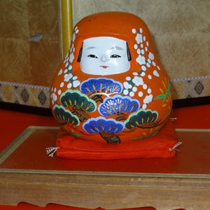 以下が含まれることがあります： 伝統的な日本のダルマ人形。オレンジ色で、白い花柄と青い松のデザイン。人形は赤いクッションの上に置かれ、木製のディスプレイスタンドに飾られています。背景は金色のスクリーンです。