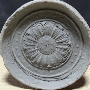 以下が含まれることがあります： 中央に蓮の花のレリーフが施され、その周囲を小さな隆起したドットの輪が囲む、円形の灰色の粘土製屋根瓦。この物体は厚く盛り上がった外縁を持ち、表面にはひび割れが見られるマットで風化した質感の仕上げが施されています。