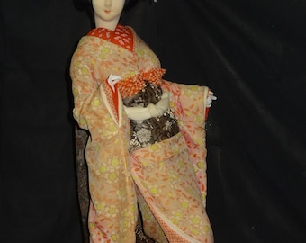 Puede incluir: Una muñeca decorativa con rostro pálido y cabello negro recogido en un moño. La muñeca viste un kimono largo de color claro con un estampado floral en tonos melocotón, amarillo y verde, sobre un obi marrón oscuro con una faja blanca.