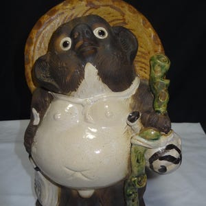 May include: A ceramic Tanuki statue, a traditional Japanese raccoon dog figure. It has a large belly, a straw hat, and holds a sake bottle and a staff. The statue is primarily brown, cream, and green.