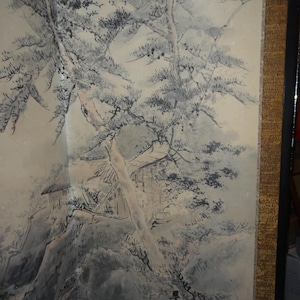 May include: A detailed ink wash painting on a folding screen. The artwork depicts a mountainous landscape with trees, a building, and a stream. The color palette is primarily black, white, and gray, with a gold border.