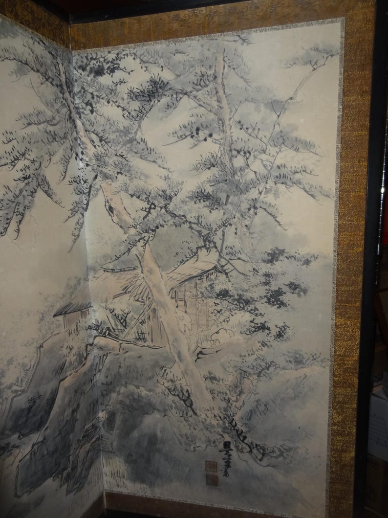 May include: A detailed ink wash painting on a folding screen. The artwork depicts a mountainous landscape with trees, a building, and a stream. The color palette is primarily black, white, and gray, with a gold border.