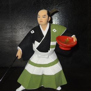 Puede incluir: Figura de un samurái japonés con vestimenta tradicional. La figura lleva un kimono negro y blanco con detalles verdes y una falda blanca. Sostiene un cuenco rojo y una lanza negra.