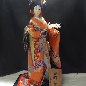 Puede incluir: Muñeca japonesa con kimono tradicional, con estampados florales naranjas, rojos y morados. La muñeca tiene una expresión serena, cabello negro y accesorios florales. Un letrero de madera con caracteres japoneses está en la base.