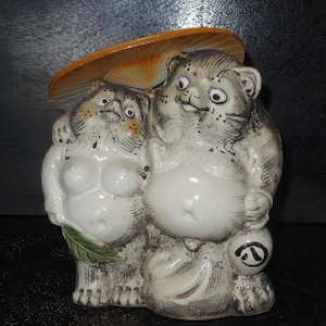 May include: A ceramic Tanuki statue depicting two raccoon dogs. The figures are white with gray and brown details, wearing a straw hat. One holds a sake bottle. The statue has a whimsical, traditional Japanese aesthetic.
