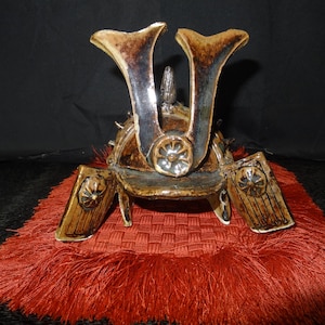 Puede incluir: Casco de samurái de cerámica en marrón y dorado, con crestas distintivas en forma de ala. Descansa sobre una base de tela roja texturizada, sobre un fondo oscuro. El diseño del casco incluye detalles intrincados y una pieza frontal decorativa.