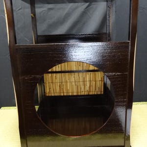 以下が含まれることがあります： 濃い茶色の漆塗りの木製棚ユニット。正方形のフレームの前面パネルには大きな円形の切り抜きが施されています。内側の背面壁には明るい黄褐色の竹のすだれが張られており、光沢のある暗い木材の仕上げと対照的な質感を生み出しています。