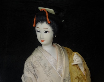 Pode incluir: Uma boneca gueixa japonesa com tez pálida, cabelo preto e detalhes em vermelho. A boneca usa um quimono creme com estampas florais e uma camada externa amarela. O rosto da boneca tem traços delicados e lábios vermelhos.