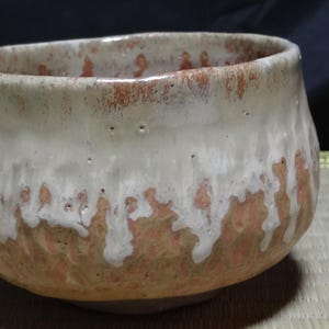 May include: A ceramic tea bowl with a rounded shape, featuring a textured terracotta-coloured base and a thick, white glaze that drips down from the rim. The surface shows visible variations in colour and texture, with a wide, open mouth.