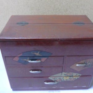 May include: A small, red wooden box with four drawers. The drawers have decorative designs of fans and birds. The box has a hinged lid.