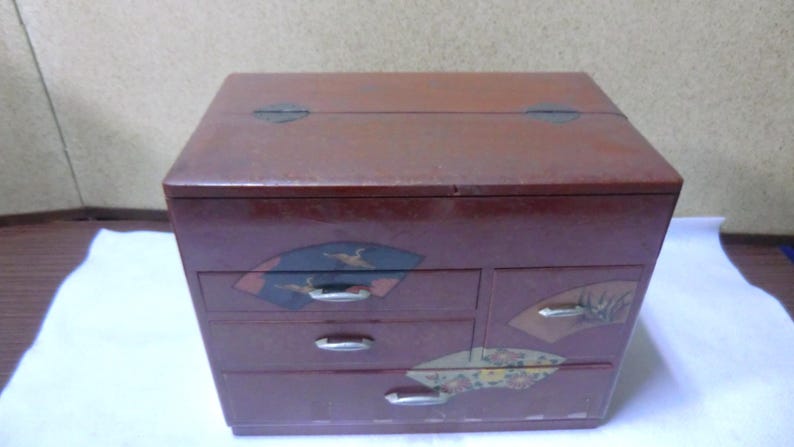 May include: A small, red wooden box with four drawers. The drawers have decorative designs of fans and birds. The box has a hinged lid.