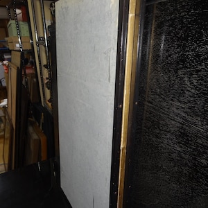 May include: A large, rectangular panel with a light gray surface, framed in dark brown wood with gold accents. The panel appears to be part of a larger piece, possibly a screen or room divider. The background is cluttered.