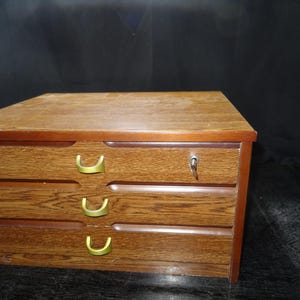 Puede incluir: Un mueble de madera de tres cajones con cerradura y llave. El mueble tiene forma rectangular con una tapa marrón claro y un marco marrón más oscuro. Los cajones tienen tiradores de color latón.