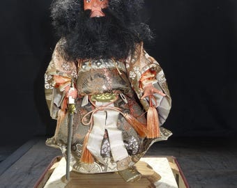 May include: A Japanese doll of a warrior figure, featuring a stern expression and a long black beard. The doll wears a detailed kimono with gold and brown patterns, holding a sword. It stands on a wooden base with a decorative tray.