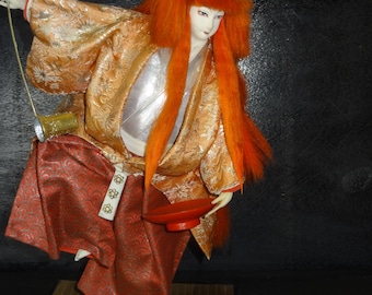 Puede incluir: Muñeca japonesa con una larga peluca naranja, vestida con un kimono dorado y rojo estampado. La muñeca sostiene un arco y un pequeño plato rojo. Está sobre una base de madera.