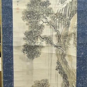 May include: A vertical hanging scroll featuring an ink wash painting of a waterfall cascading down a rocky cliffside with pine trees and a small figure crossing a stone bridge. The artwork is framed by a dark blue textured fabric border.