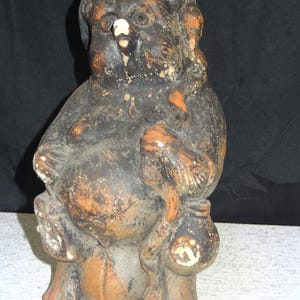 Peut inclure: Statue de Tanuki en céramique patinée. La figurine est principalement noire avec des accents orange et blancs. Elle a un gros ventre, tient une bouteille de saké et un sac, et porte un chapeau. La statue a un aspect vieilli et usé.