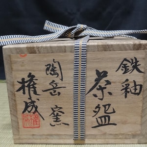 May include: A rectangular wooden box with a light brown grain, tied with a blue and white striped fabric cord. The front of the box features black Japanese calligraphy characters and a small red square stamp, indicating its contents and origin.