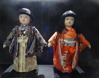 May include: Two Japanese dolls are displayed. The doll on the left wears a blue and gold patterned kimono with a black hat. The doll on the right wears a red and orange kimono with long black hair. Both dolls have pale skin and are standing on black bases.
