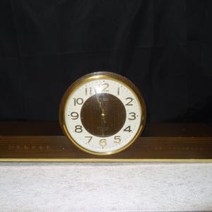 Op de afbeelding: Een vintage houten klok met een ronde wijzerplaat en gouden rand. De wijzerplaat heeft zwarte cijfers op een witte achtergrond. De klok staat op een rechthoekige houten basis. De wijzers van de klok wijzen naar 1:00 uur.