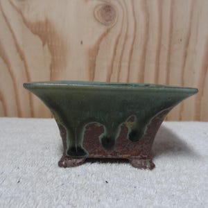 Könnte beinhalten: Ein quadratischer Bonsai-Topf aus Keramik mit grüner Glasur und braunen Akzenten. Der Topf hat einen Tropf-Glazureffekt und vier kleine Füße.