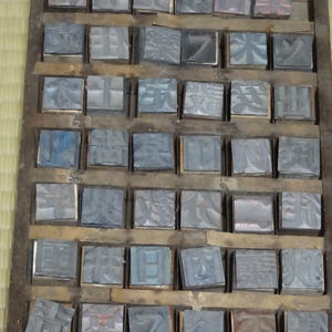 May include: A rectangular wooden tray containing a grid of individual metal printing blocks. Each block features a unique raised Japanese character or symbol on its top surface. The blocks are arranged in rows and columns within the wooden frame.