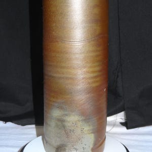 May include: A cylindrical, handmade ceramic vase with a gradient of colors. The top is a warm brown, transitioning to a lighter, almost metallic gray at the base. The vase has a textured surface with horizontal lines.