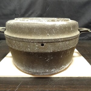 May include: A dark gray, cast iron cooking pot with a lid and side handles. The pot sits on a light-colored, triangular trivet. The pot has a circular shape and appears to be vintage.