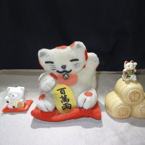 以下が含まれることがあります： 日本の招き猫の置物のコレクション。一番大きいものは白と赤のアクセントで、金のコインを持ち、赤いクッションの上に座っています。小さな猫が赤いマットの上に座っています。