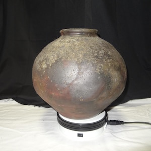 Könnte beinhalten: Ein großer, runder Steinguttopf mit dunkler, strukturierter Oberfläche. Der Topf hat einen breiten Körper und einen schmalen Hals. Die Farben reichen von Braun bis Schwarz, mit helleren Bereichen in Beige. Er steht auf einer weißen Drehplattform.