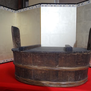 May include: A weathered, wooden Japanese bath tub, or ofuro, with a dark finish. The tub has a rectangular shape with rounded sides, a woven band around the middle, and two vertical handles. The lid is made of dark wood.