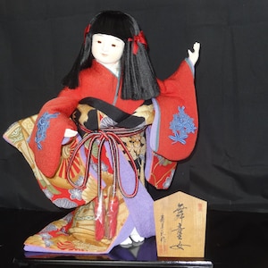 Puede incluir: Muñeca japonesa con kimono rojo, pelo negro y letrero de madera. El kimono presenta estampados florales y detalles azules. La muñeca posa con un brazo levantado, el letrero tiene caracteres japoneses.