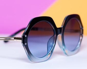 May include: Oversized black and blue gradient sunglasses with a hexagonal frame. The lenses transition from brown to blue, and the arms have gold accents. The sunglasses are set against a colorful background.