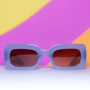 May include: A pair of lavender-colored sunglasses with rectangular frames and brown lenses. The sunglasses are set against a colorful background of yellow, pink, and orange.