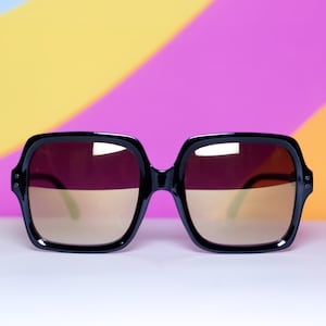 May include: Black square-framed sunglasses with gradient brown lenses. The sunglasses have a thick frame and a retro aesthetic. The lenses transition from a dark brown at the top to a lighter brown at the bottom. The background is a colorful blend of yellow, pink, and purple.