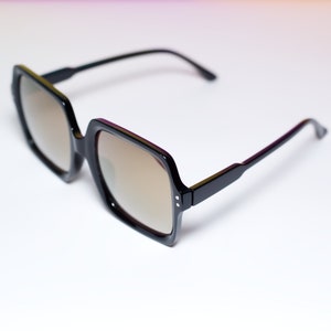 May include: Black square-framed sunglasses with brown tinted lenses. The sunglasses have a thick frame and sleek black arms. The lenses are a light brown color, and the overall design is modern and stylish.