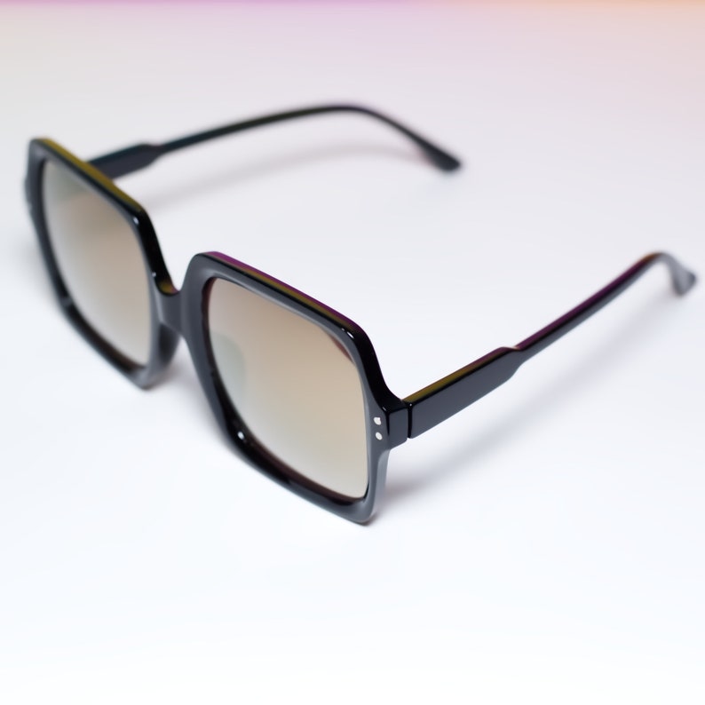 May include: Black square-framed sunglasses with brown tinted lenses. The sunglasses have a thick frame and sleek black arms. The lenses are a light brown color, and the overall design is modern and stylish.
