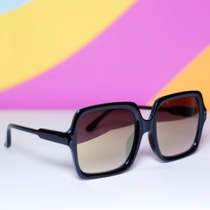 May include: Black square-framed sunglasses with gradient brown lenses. The sunglasses have a thick frame and a classic design. The background is a colorful blend of yellow, pink, and purple.