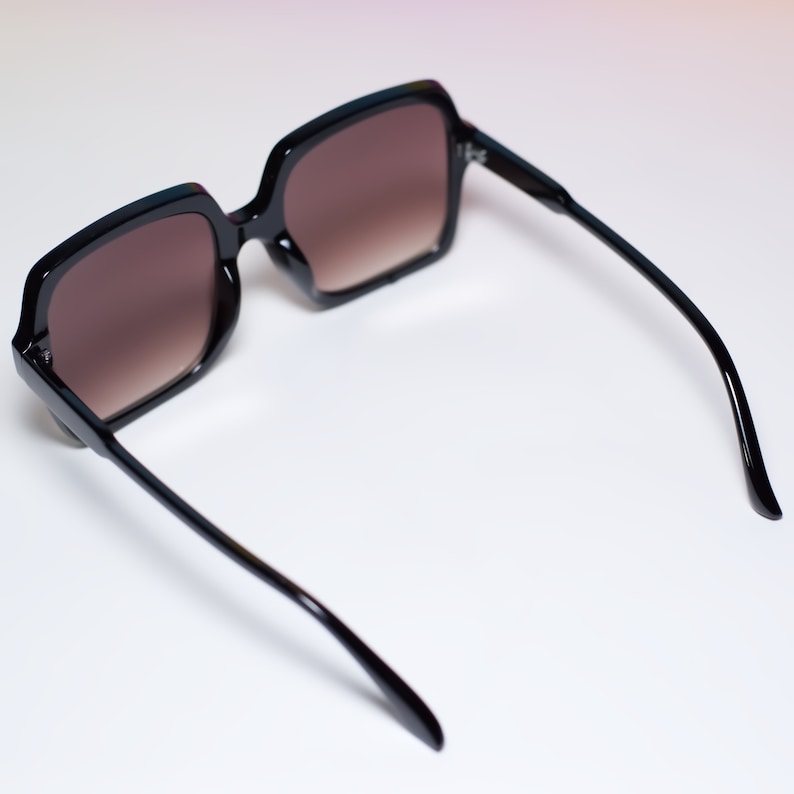 May include: Black square-framed sunglasses with gradient brown lenses. The sunglasses have a thick frame and sleek, curved arms. The lenses transition from a darker shade at the top to a lighter shade at the bottom.